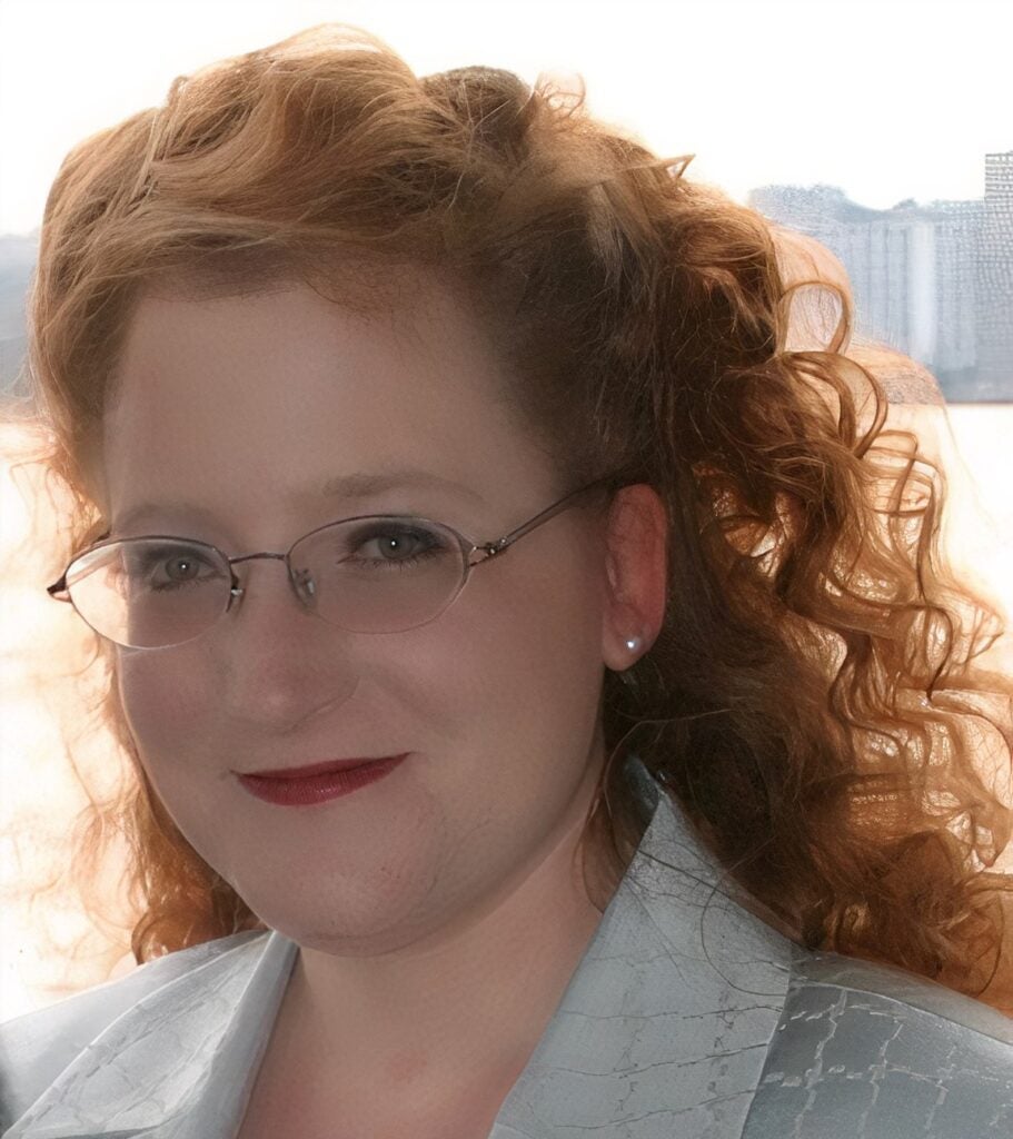 A picture of Morgan Whitlatch, a white woman with red curly hair, who is wearing glasses and a light blue suit and smiling at the camera.