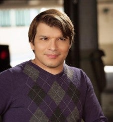 Andy is pictured smiling in a headshot with a blurred background. He is a Latin X man with an olive complexion, short brown highlighted hair, and brown eyes, and he is wearing a purple argyle sweater. You can see the top of his wheelchair in the photo.