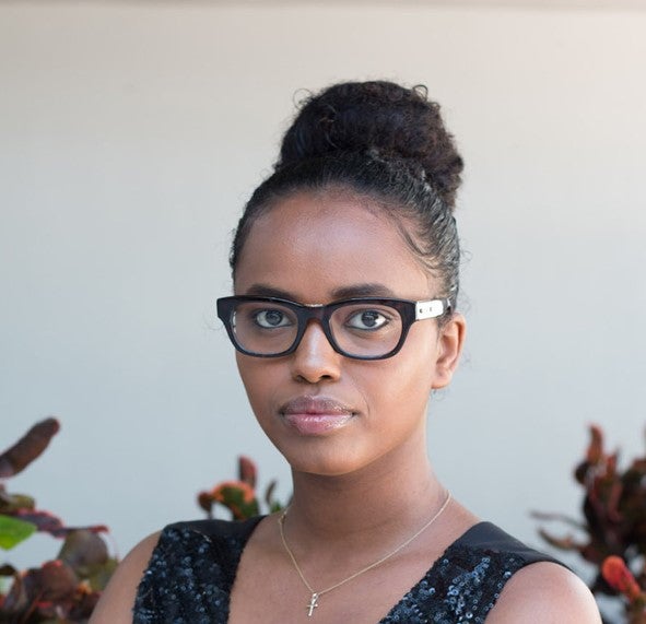 Betelhem is pictured in a headshot with a light gray background and some plants at the bottom. She is a black woman with a brown complexion, curly black hair tied in a top knot, dark brown eyes, and is wearing a black sequin dress, glasses, and a gold necklace.