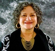 A smiling picture of Diane, an older white woman with graying short curly care, a black and white sweater and hoop earrings.