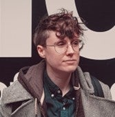 A headshot of Elio, a white person with messy short brown hair and glasses, gaze directed to the right, standing in front of an indistinct black and white graphic.