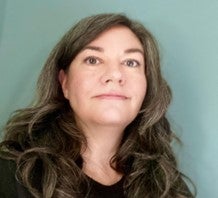 Ellen is a pinkish, white woman with long and wavy dark hair with white and gray streaks and brown eyes.  Ellen is wearing a black shirt and standing in front of a light blue wall.