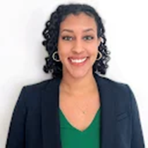 Kalie is pictured smiling in a headshot with a light-gray background. She is a biracial, Black-American woman with an olive complexion, shoulder-length, curly, black hair, brown eyes, and is wearing a green blouse with a navy blazer and gold hoop earrings.