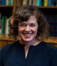 A headshot of Libbie, a middle-aged white woman with curly, light brown chin-length hair, and a blue blouse, open at the neck. She sits in front of a partially full bookshelf, smiling. 