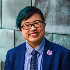 Photo by Sarah Tundermann: Lydia X. Z. Brown smiles in front of reeds blowing in a gentle breeze. They are an East Asian nonbinary/transmasc person with short black hair and glasses, wearing a dark blue suit and blue paisley necktie.