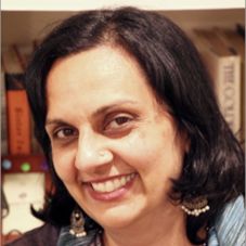 A headshot of Pamala, a middle-aged brown woman with short black hair and brown eyes. Pamala is looking at the camera and smiling in this picture and wearing silver beaded earrings and a green and gray tunic. She is pictured in front of a bookcase in her home.