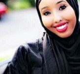 A headshot of Safiya, a black woman wearing a black covered hijab (headscarf), and a black colored long sleeve dress. She is seen standing outside of her neighborhood– smiling and posing for the camera. 