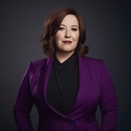 Professional portrait of a woman. She has shoulder-length brown hair styled in loose waves. Her makeup includes dark eyeshadow and red lipstick. She's wearing a vibrant purple blazer over a black top. The woman has a confident, friendly expression with a slight smile. The background is a dark, neutral color, creating a studio portrait effect that draws focus to the subject.