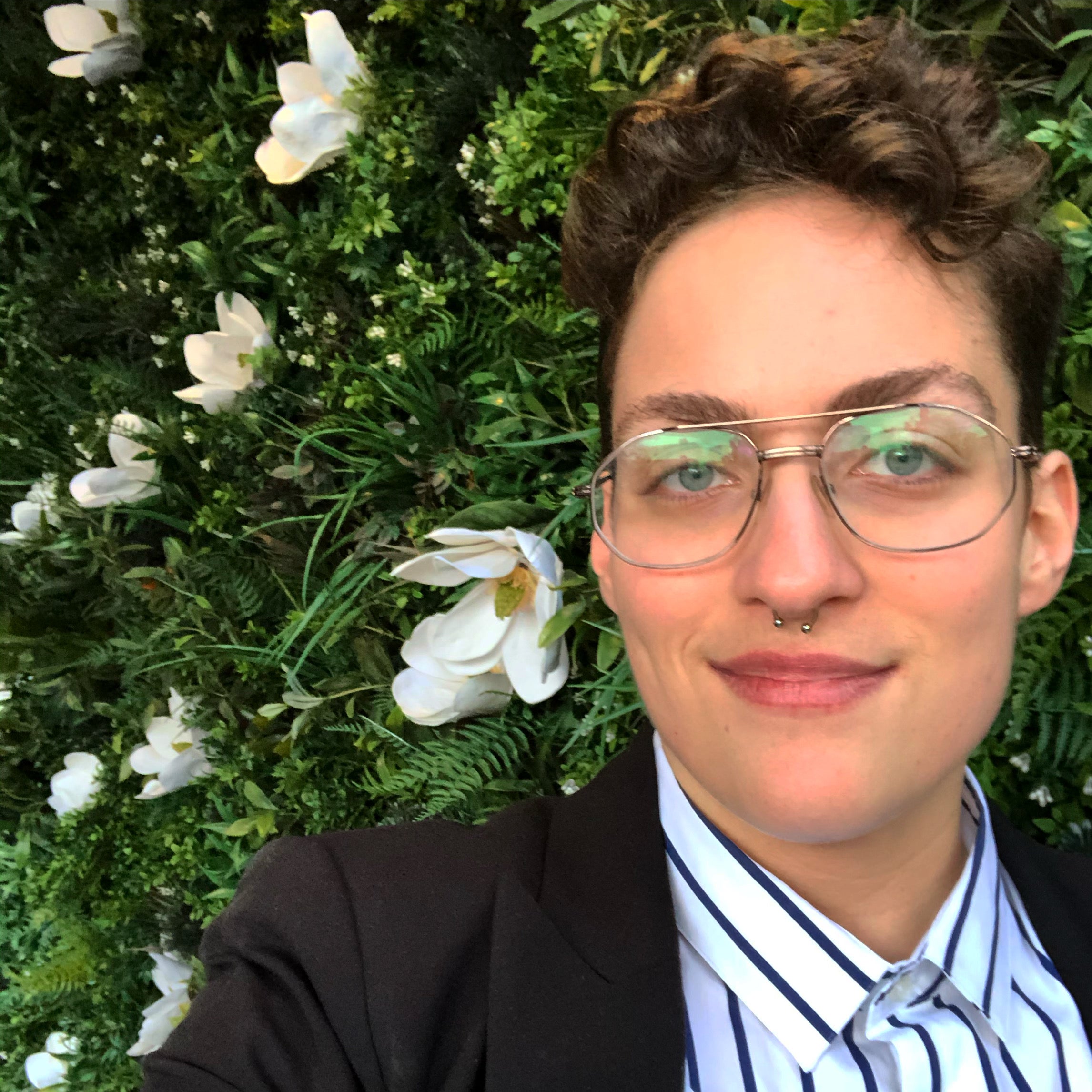 A white person with short brown curly hair wearing glasses, with a smirk on their face. They are wearing a button-up white and blue striped shirt a black sportcoat and behind them there is a bed of white flowers in green grass.