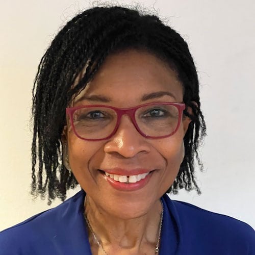 Tawara is pictured smiling, wearing a royal blue blouse, silver and blue necklace, and red eyeglasses.