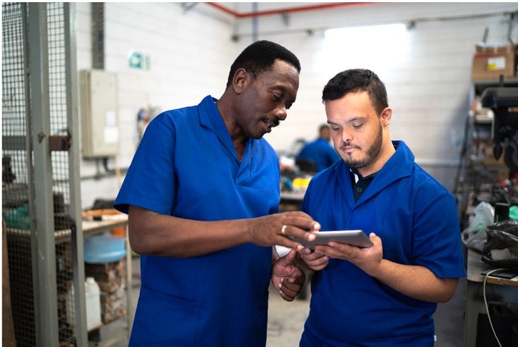 An older employee shows a younger employee with Down Syndrome something on a tablet. They are wearing uniforms and in a shop.