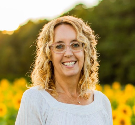 A headshot of Professor Dana Childress. Professor Childress is a middle-aged white woman with curly blonde hair and glasses. She is wearing a white shirt and is smiling. Her background, a field of yellow flowers, is blurred.