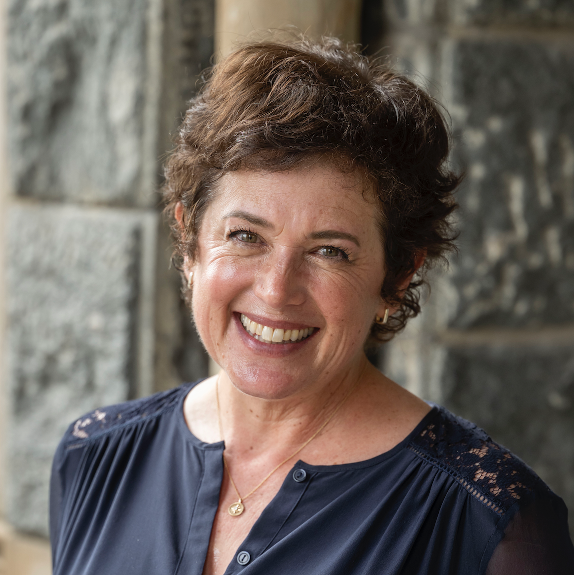 A headshot of a smiling woman with short, curly brown hair, wearing a navy blue blouse with lace detailing on the shoulders and a small gold pendant necklace. She is standing in front of a textured gray stone wall.